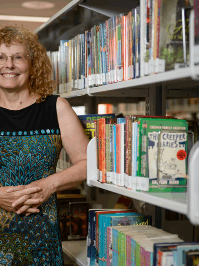 A woman in a black and blue dress smiles and stands in front of bookshelves in a public library.