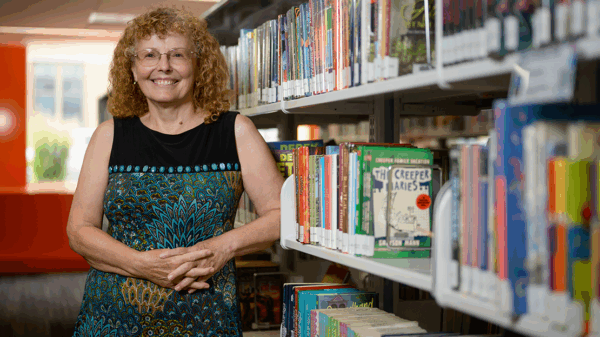 A woman in a black and blue dress smiles and stands in front of bookshelves in a public library.