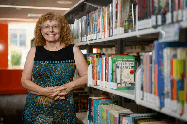 A woman in a black and blue dress smiles and stands in front of bookshelves in a public library.