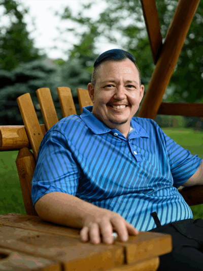 A smiling man wearing a blue shirt is pictured sitting on a wooden chair outside.