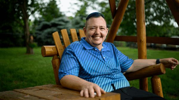 A smiling man wearing a blue shirt is pictured sitting on a wooden chair outside.