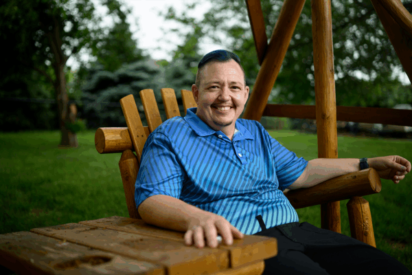 A smiling man wearing a blue shirt is pictured sitting on a wooden chair outside.